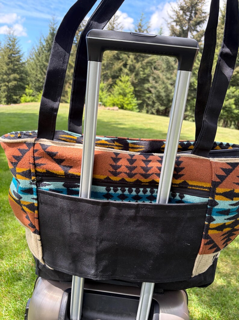 May include: A patterned tote bag with black straps and a black pocket, attached to a rolling suitcase. The bag features a colourful geometric design in shades of brown, orange, blue, and cream. The background shows a green lawn and trees.