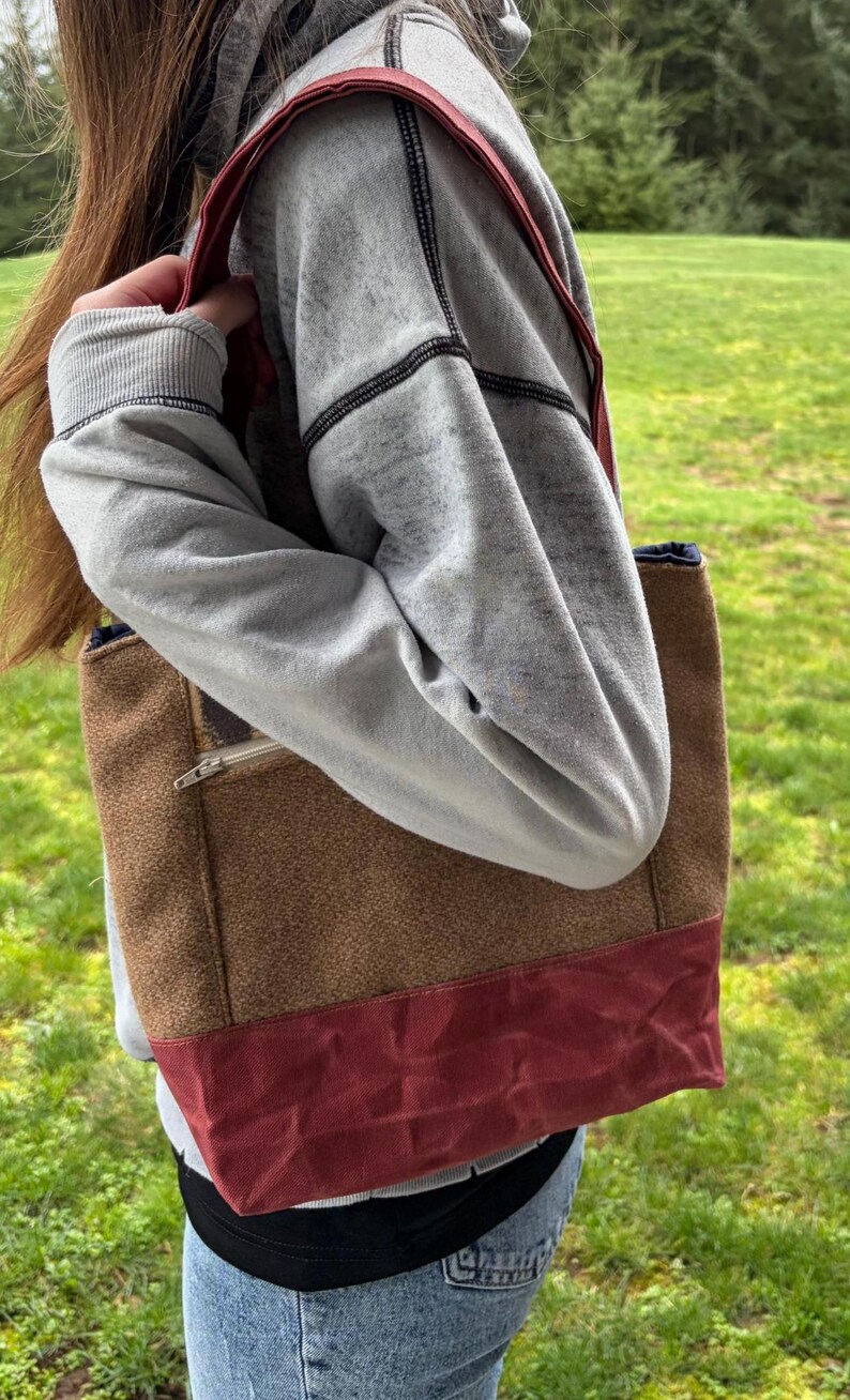 May include: A brown and red tote bag with a zipper pocket. The bag has a red strap and base, with a brown textured body. The bag is being carried over the shoulder.