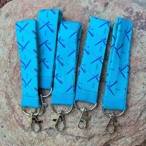May include: Five turquoise wrist keychains with a dragonfly pattern in dark blue and red. Each keychain has a silver-colored clasp. The keychains are arranged on a textured, neutral-colored surface.