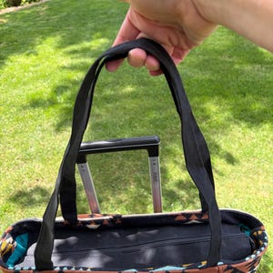 May include: A patterned tote bag with black handles is held up against a green grassy background. The bag features a geometric design in shades of beige, brown, and black. The bag has a black zip closure.