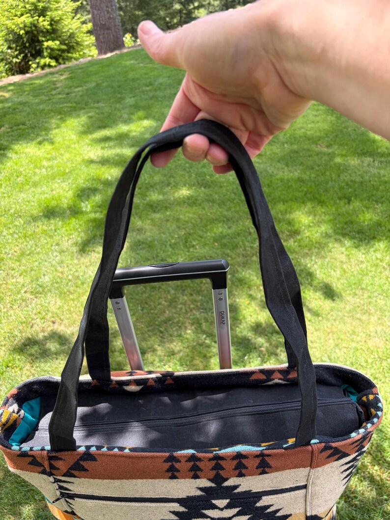 May include: A patterned tote bag with black handles is held up against a green grassy background. The bag features a geometric design in shades of beige, brown, and black. The bag has a black zip closure.