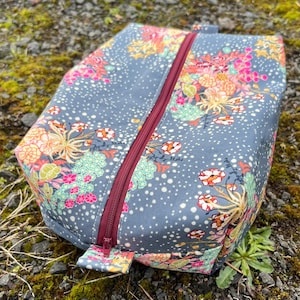 May include: A gray zippered pouch with a floral print. The pouch has a red zipper and a white polka dot pattern.
