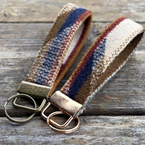 May include: Two keychains with fabric straps. The straps feature a plaid pattern in shades of brown, blue, and cream. Each keychain has a metal clasp and a ring for keys. The keychains are displayed on a wooden surface.