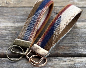 May include: Two keychains with fabric straps. The straps feature a plaid pattern in shades of brown, blue, and cream. Each keychain has a metal clasp and a ring for keys. The keychains are displayed on a wooden surface.