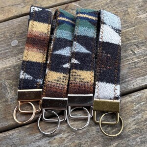 May include: Four keychains with patterned fabric straps in shades of brown, tan, blue, and white. Each keychain has a metal clasp and a split ring. The keychains are displayed on a wooden surface.
