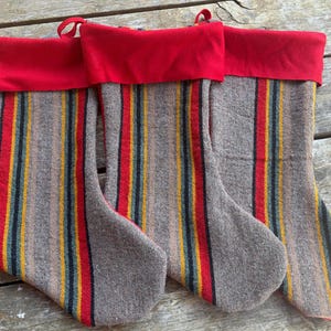 May include: Three Christmas stockings featuring a textured grey base with vertical stripes in red, yellow, and black. Each stocking has a solid red cuff at the top and a small red fabric loop for hanging. The stockings are arranged side by side.