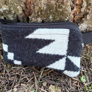 May include: A small, rectangular black and white patterned pouch with a zipper closure and a small black wrist strap. The pouch features a geometric design and a gold-colored metal clasp.