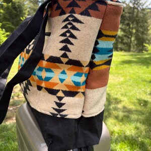 May include: A large tote bag with a Southwestern-inspired pattern in beige, black, brown, orange, and turquoise. The bag has black straps and a black base. It is sitting on top of a silver suitcase, outdoors.