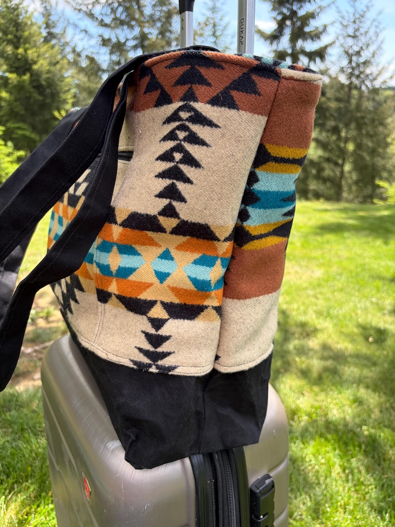 May include: A large tote bag with a Southwestern-inspired pattern in beige, black, brown, orange, and turquoise. The bag has black straps and a black base. It is sitting on top of a silver suitcase, outdoors.