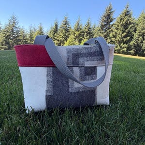 May include: A tote bag featuring a red top panel, a white textured lower section, and a gray geometric pattern on the side. The bag includes two gray fabric shoulder straps attached to the top, with a structured rectangular shape and visible stitching.