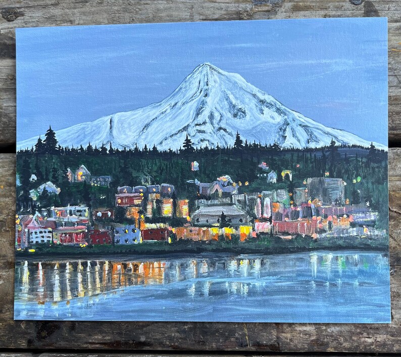 May include: A painting of a snow-capped mountain with a town in the foreground. The town is lit up with lights and there is a body of water in front of it. The sky is a light blue color.