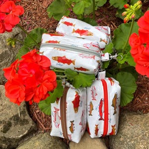 May include: White zippered pouches with a repeating pattern of red, orange, and brown fish. The pouches have a silver zipper and a small wrist strap. The pouches are surrounded by red flowers and green leaves.