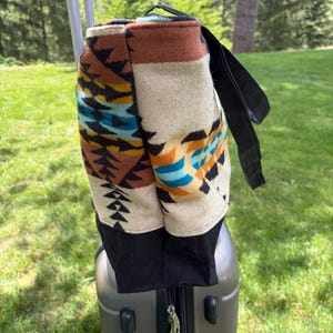 May include: A patterned tote bag with a black base and strap rests on a grey suitcase. The bag features a geometric design in shades of brown, beige, teal, and black. The background shows a green lawn and trees.