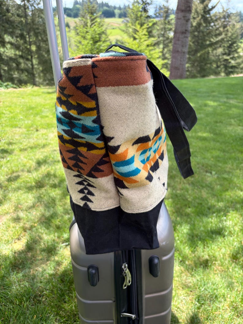 May include: A patterned tote bag with a black base and strap rests on a grey suitcase. The bag features a geometric design in shades of brown, beige, teal, and black. The background shows a green lawn and trees.