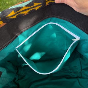 May include: Open tote bag with a black exterior and a turquoise interior. The bag features a white zip and a patterned top with yellow, orange, and turquoise geometric designs. The interior has a pocket with a white trim.