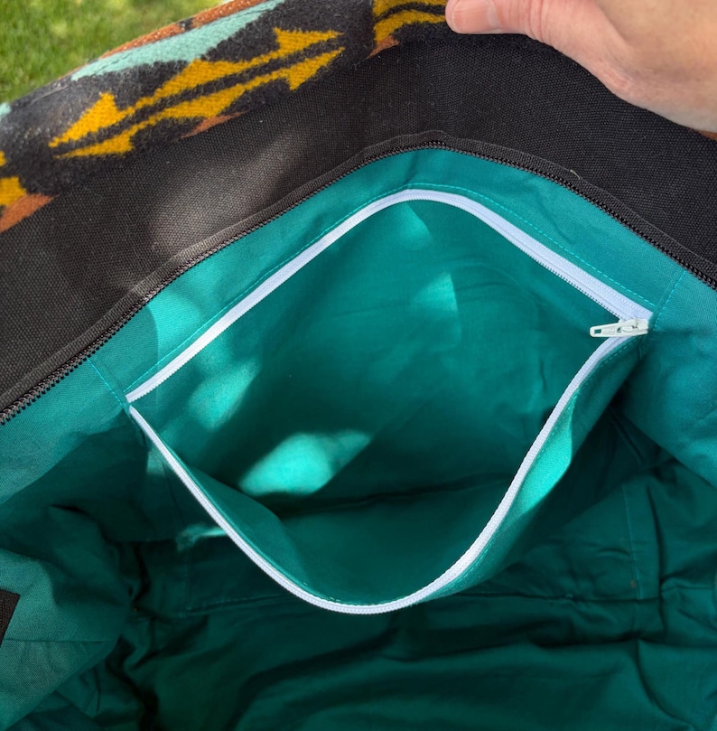 May include: Open tote bag with a black exterior and a turquoise interior. The bag features a white zip and a patterned top with yellow, orange, and turquoise geometric designs. The interior has a pocket with a white trim.