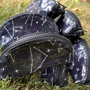 May include: A collection of black zippered pouches with a constellation pattern in white. The pouches are in various shapes and sizes, with a shiny, clear finish. The zippers are black.