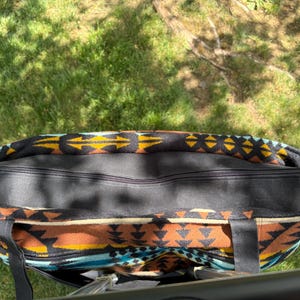 May include: A large tote bag featuring a Southwestern pattern in black, brown, yellow, and turquoise. The bag has a black zip closure and black handles. The bag is on green grass.