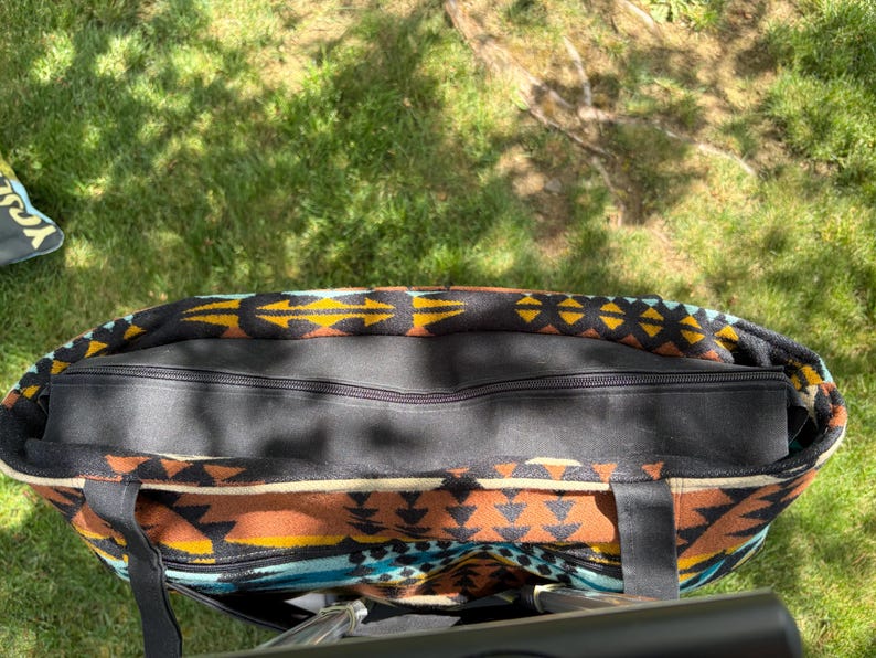 May include: A large tote bag featuring a Southwestern pattern in black, brown, yellow, and turquoise. The bag has a black zip closure and black handles. The bag is on green grass.