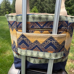 May include: A patterned tote bag with a beige handle and a zipper pocket. The bag features a geometric design in shades of blue, yellow, and green. The bag is attached to a silver suitcase handle.