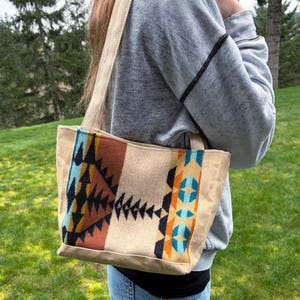 May include: A beige tote bag with a colorful geometric pattern in shades of blue, orange, and brown. The bag has a canvas-like texture and a matching beige shoulder strap. The bag is carried over the shoulder.