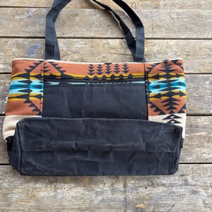 May include: A black canvas tote bag with a Southwestern-inspired patterned design. The bag has a zippered pocket and strong black handles. The patterned section includes shades of brown, blue, yellow, and cream.