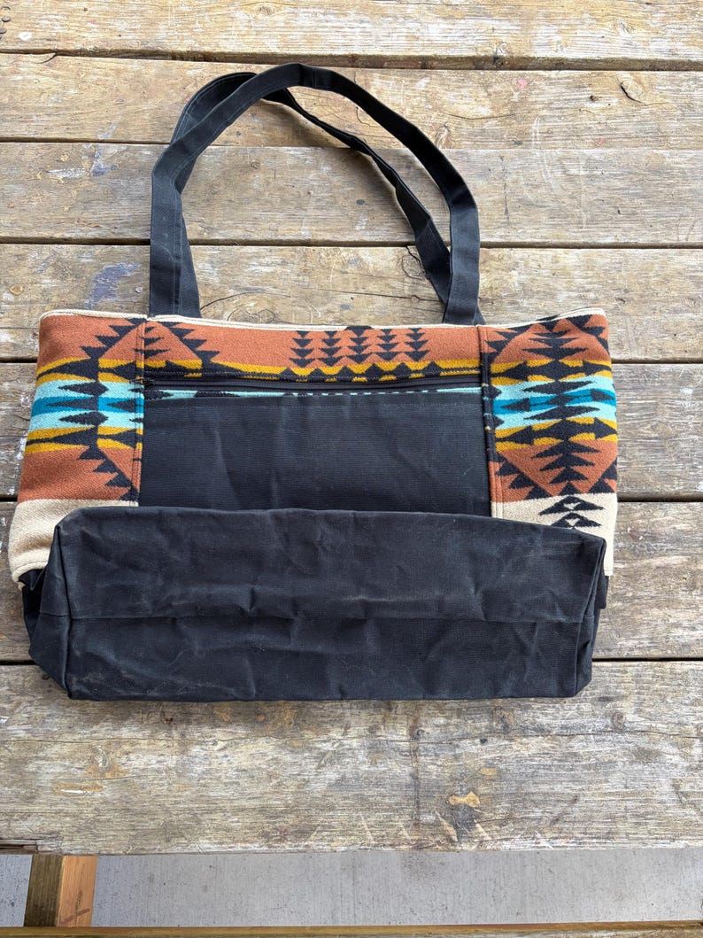 May include: A black canvas tote bag with a Southwestern-inspired patterned design. The bag has a zippered pocket and strong black handles. The patterned section includes shades of brown, blue, yellow, and cream.