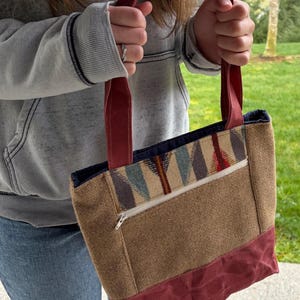 May include: A brown tote bag with burgundy accents and handles. The bag features a patterned design on the upper portion and a zippered pocket. The bottom and handles are a solid burgundy color.