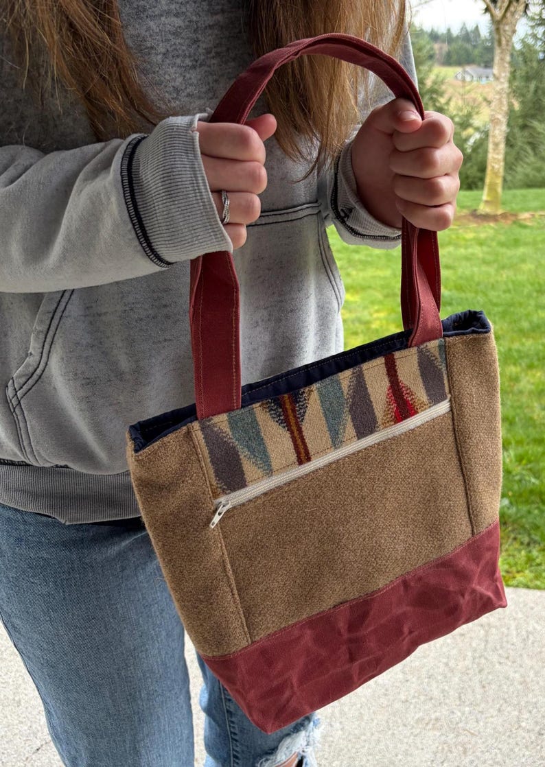 May include: A brown tote bag with burgundy accents and handles. The bag features a patterned design on the upper portion and a zippered pocket. The bottom and handles are a solid burgundy color.