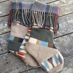 May include: Two holiday stockings crafted from a patchwork of various fabrics including beige fleece, olive green felt, and patterned woven textiles featuring blue, red, and brown geometric designs. Each stocking features a fringed cuff at the top.