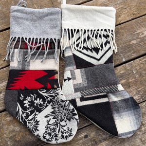 May include: Two holiday stockings made from patchwork fabric. The left stocking features a gray cuff with fringe, a red and black geometric section, and a black and white floral pattern. The right stocking has a cream cuff with fringe and black, white, and gray geometric patterns.