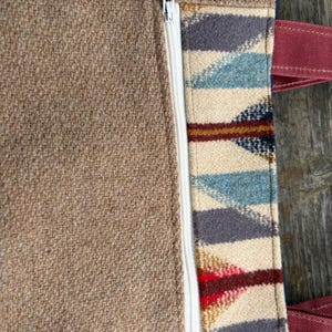 May include: Close-up of a brown tote bag with a white zipper and patterned fabric detail. The patterned section features blue, gray, red, and cream-colored geometric shapes. The bag has burgundy handles.