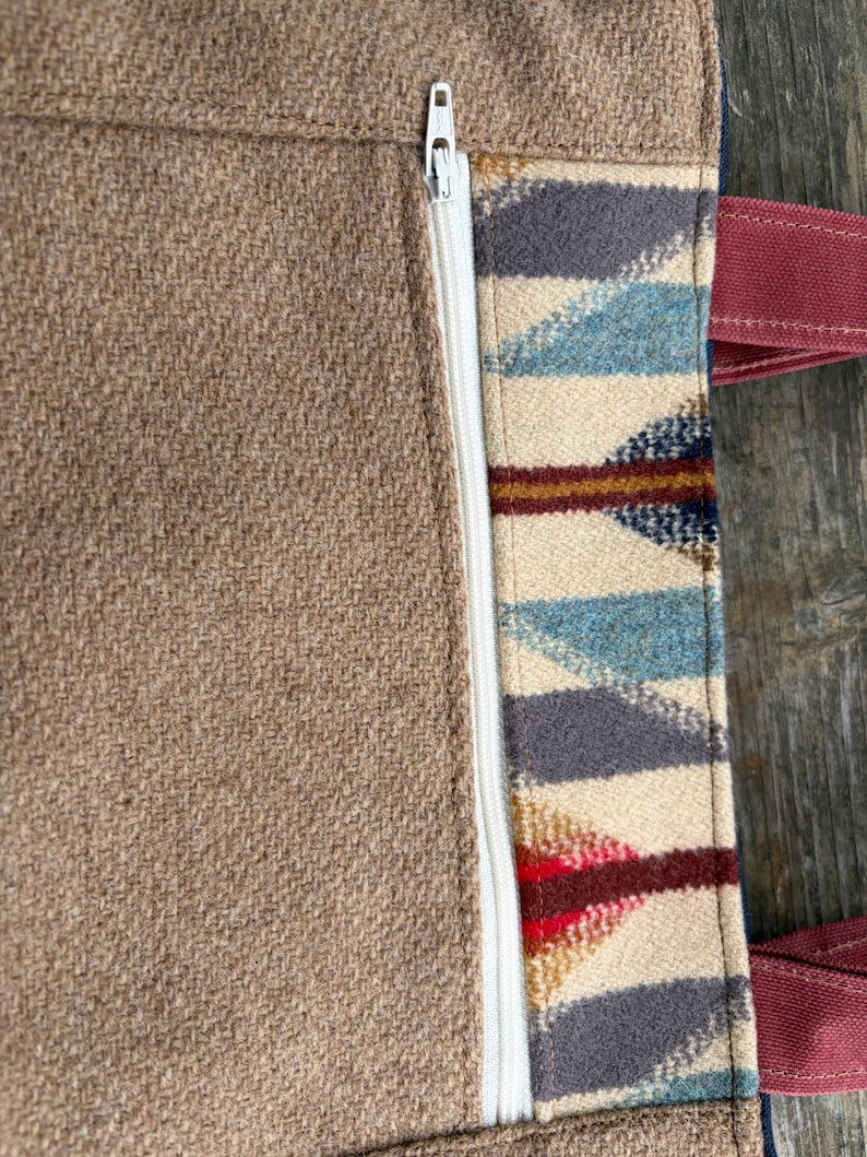 May include: Close-up of a brown tote bag with a white zipper and patterned fabric detail. The patterned section features blue, gray, red, and cream-colored geometric shapes. The bag has burgundy handles.