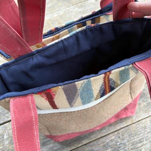 May include: A tote bag with a patterned front pocket and red straps. The bag has a tan body with a red base and straps. The interior is navy blue. The front pocket has a colorful geometric pattern with a white zipper.