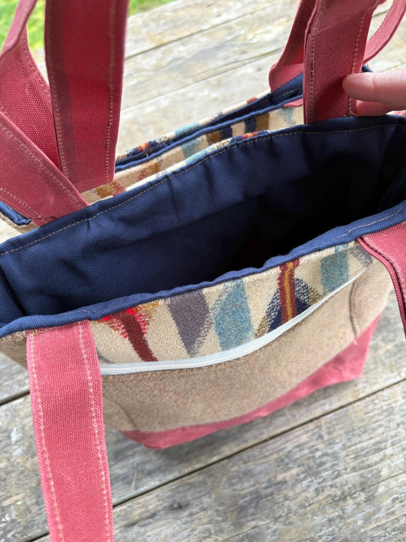 May include: A tote bag with a patterned front pocket and red straps. The bag has a tan body with a red base and straps. The interior is navy blue. The front pocket has a colorful geometric pattern with a white zipper.