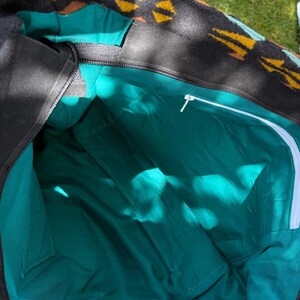 May include: An open black tote bag with a teal interior. The top edge has a patterned design in yellow, brown, and black. The interior features multiple pockets and a white zip. The bag is open, showing its capacity.