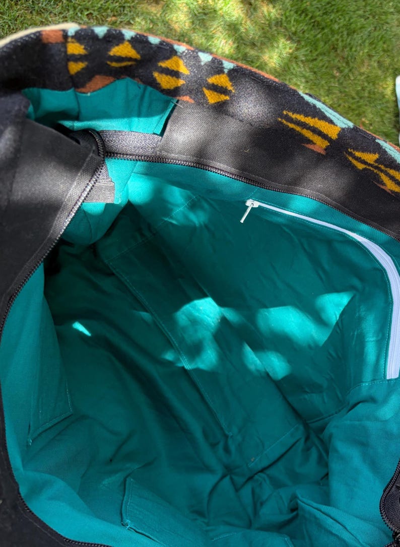 May include: An open black tote bag with a teal interior. The top edge has a patterned design in yellow, brown, and black. The interior features multiple pockets and a white zip. The bag is open, showing its capacity.