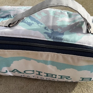 May include: A rectangular Glacier National Park lunch bag with a gray handle and a navy blue zipper. The bag features a white background with a blue and gray mountain design and the park's name in green.