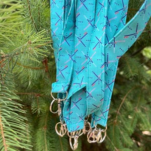 May include: A collection of teal lanyards with a repeating geometric pattern in pink, blue, and white. Each lanyard has a silver-colored swivel clasp. The lanyards are hanging in front of a backdrop of green pine needles.