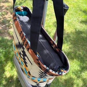 May include: A patterned tote bag with black straps and a zip closure. The bag features a geometric design in shades of brown, beige, black, and turquoise. The bag is sitting on top of a grey suitcase, with green grass in the background.