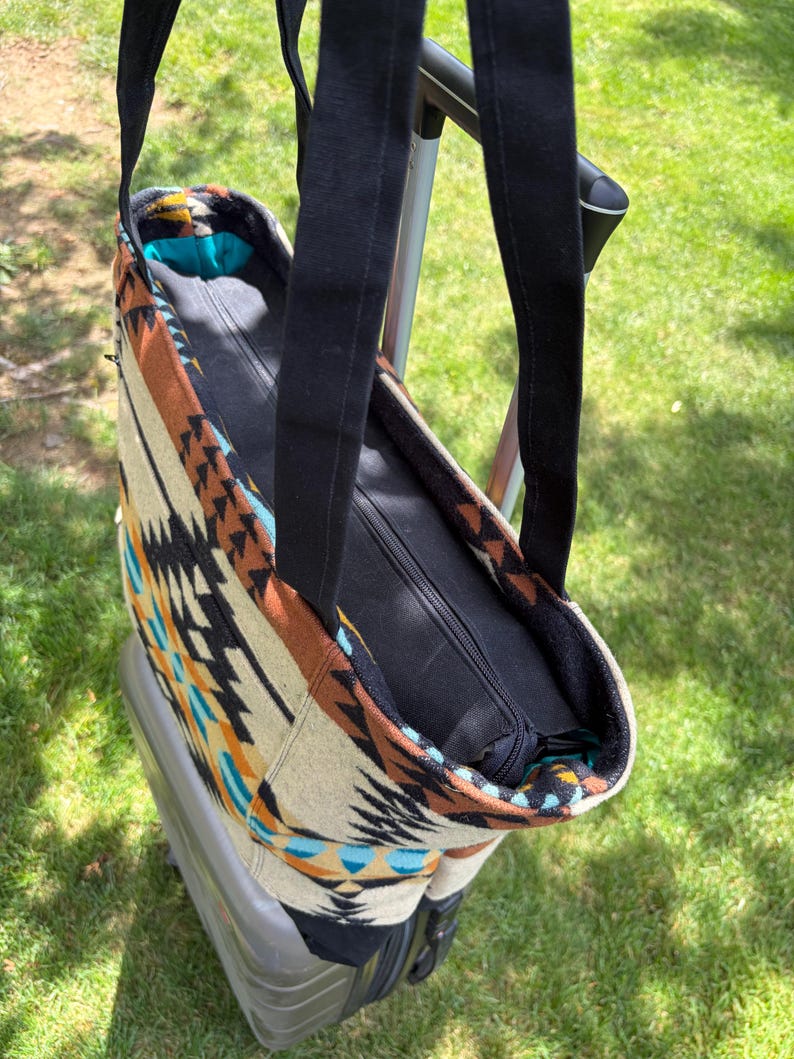 May include: A patterned tote bag with black straps and a zip closure. The bag features a geometric design in shades of brown, beige, black, and turquoise. The bag is sitting on top of a grey suitcase, with green grass in the background.