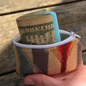 May include: A small, rectangular coin purse with a white zipper and a colorful striped pattern in shades of brown, tan, and red. The purse is open, revealing a roll of US dollar bills and a light blue object.