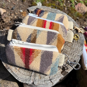 May include: Four small, rectangular zippered pouches with a geometric pattern in beige, brown, red, and blue. Each pouch has a white zipper and a small fabric loop. One pouch has a silver clasp attached.