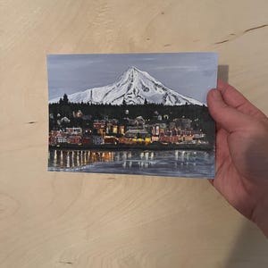 May include: A painting of a snow-capped mountain with a city in the foreground. The city is lit up with lights, and the mountain is reflected in the water in the foreground.