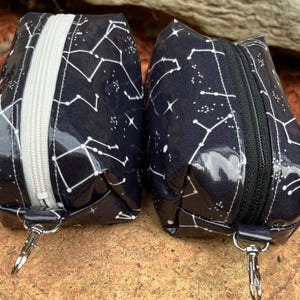 May include: Two small, dark blue pouches with a constellation pattern in white. Each pouch has a zipper closure and a silver-colored metal clasp. The pouches are sitting on a natural, textured surface.