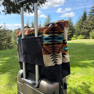 May include: A patterned tote bag with a black base sits on a silver rolling suitcase. The tote bag has a colourful geometric design in shades of brown, turquoise, and cream. The suitcase handle is extended.
