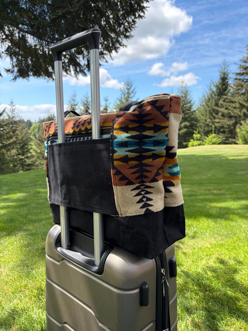 May include: A patterned tote bag with a black base sits on a silver rolling suitcase. The tote bag has a colourful geometric design in shades of brown, turquoise, and cream. The suitcase handle is extended.