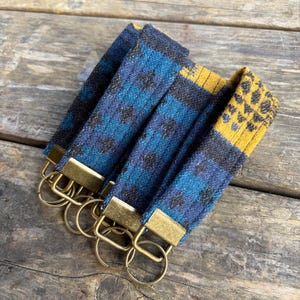 May include: A collection of key fobs with a woven fabric design in shades of blue, black, and yellow. Each fob has a brass-colored metal rectangle and a circular key ring attached.