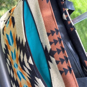 May include: A close-up of a tote bag with a Southwestern-inspired design. The bag features a colourful geometric pattern in shades of beige, orange, black, and turquoise. A turquoise interior is visible through the open zip.