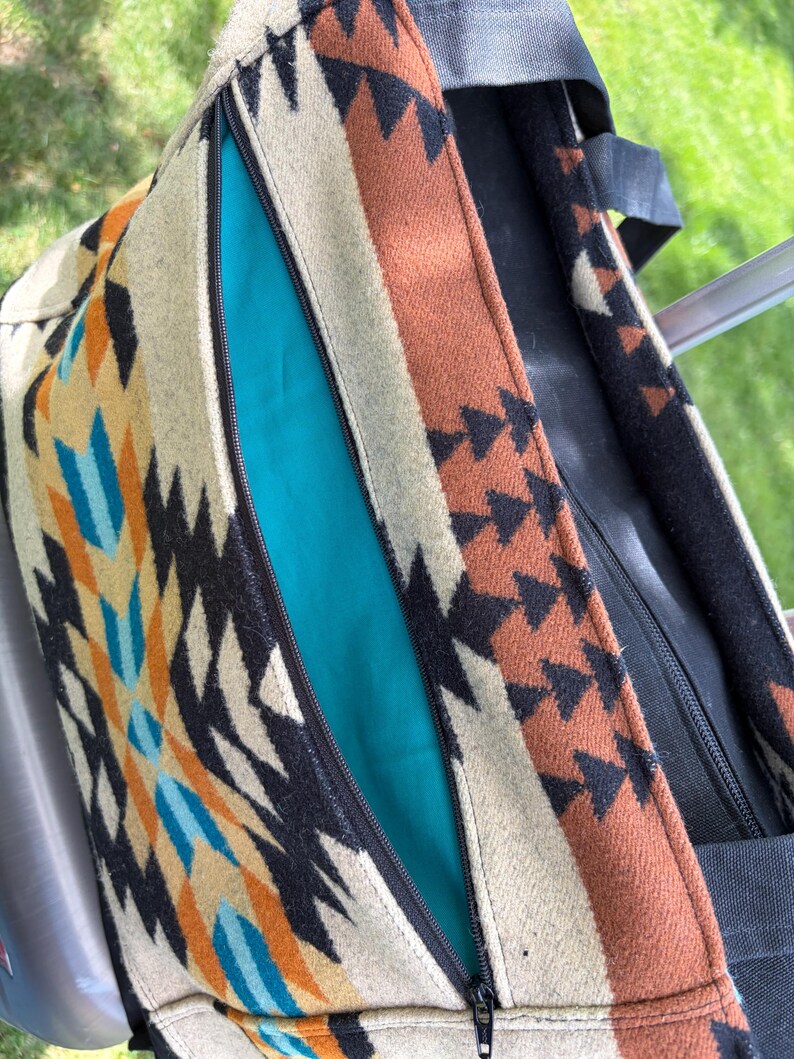 May include: A close-up of a tote bag with a Southwestern-inspired design. The bag features a colourful geometric pattern in shades of beige, orange, black, and turquoise. A turquoise interior is visible through the open zip.